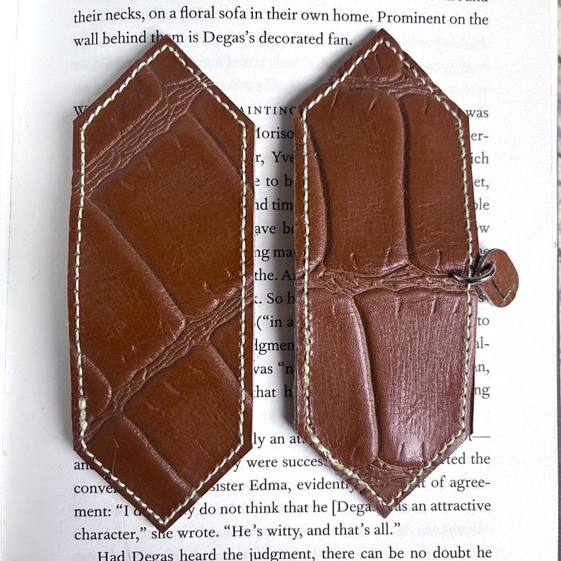 May include: Two brown leather bookmarks with a stitched edge and a crocodile pattern. The bookmarks are shaped like a pointed rectangle with rounded corners.