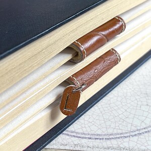 May include: Close-up of a book with two brown leather bookmarks. The top bookmark is plain, while the bottom bookmark has a metal charm with the letter "L" on it.
