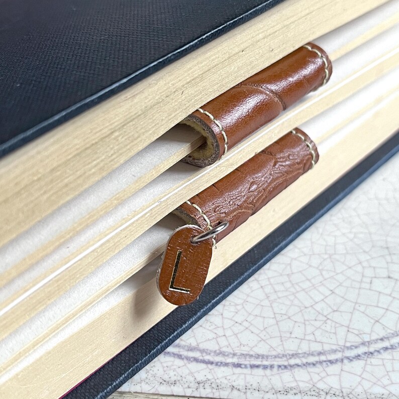 May include: Close-up of a book with two brown leather bookmarks. The top bookmark is plain, while the bottom bookmark has a metal charm with the letter "L" on it.