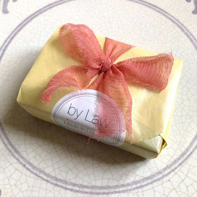May include: A yellow gift box with a pink ribbon tied in a bow. The box has a white label with the text "by Law" and "Made in London".