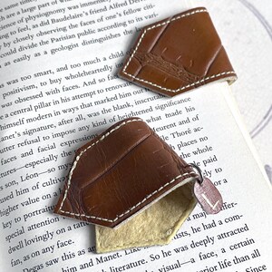 May include: Two brown leather bookmarks with a stitched edge and a cream-colored backing. The bookmarks are shaped like a pointed rectangle and have a textured surface. One bookmark has a metal charm with the letter L on it.