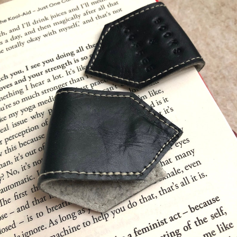 May include: Two black leather book page holders with white stitching and the word "BOOKS" embossed on one. The holders are placed on the pages of a book, with the text visible.
