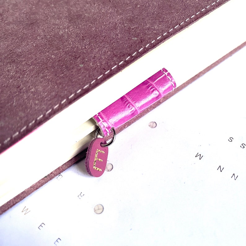 May include: A purple leather bookmark with a gold embossed letter "E" hanging from a ring. The bookmark is attached to a brown leather journal.