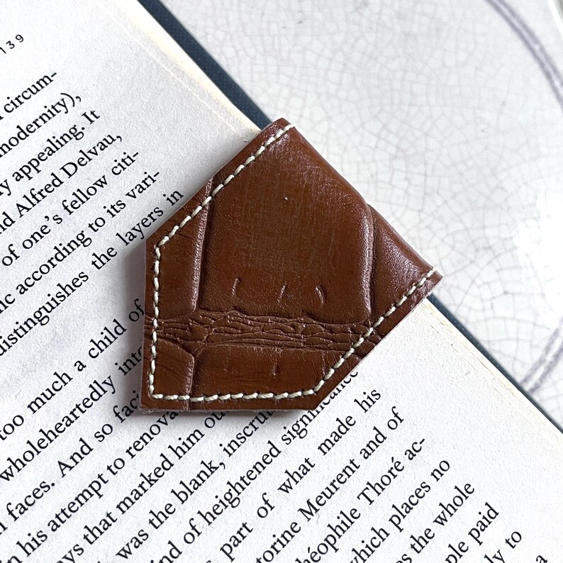 May include: A brown leather bookmark with a stitched edge and a crocodile pattern. The bookmark is folded into a triangle shape and is placed on a book.