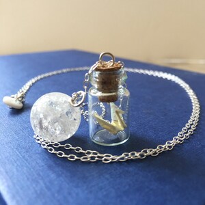 Pu&ograve; includere: Una collana con catena d'argento con un ciondolo a forma di bottiglia di vetro trasparente che contiene una gru origami bianca e un ciondolo a forma di sfera di cristallo.