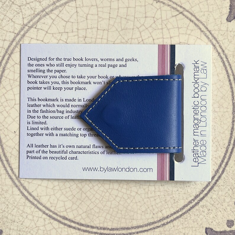 May include: A blue leather magnetic bookmark with white stitching. The bookmark is made in London by Law. The text on the card reads: "Designed for the true book lovers, worms and geeks, the ones who still enjoy turning a real page and smelling the paper. Wherever you chose to take your book or where book takes you, this bookmark won't slip, this pointer will keep your place. This bookmark is made in London, leather which would normally be used in the fashion/bag industry. Due to the source of leather, it is limited. Lined with either suede or organic leather, together with a matching top thread. All leather has it's own natural flaws and part of the beautiful characteristics of leather. Printed on recycled card. www.bylawlondon.com"