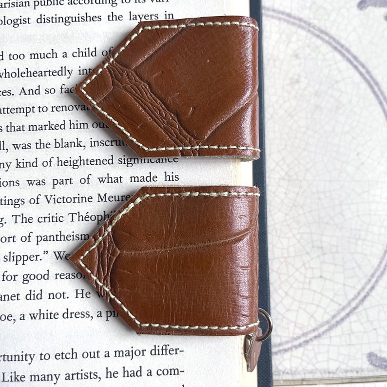 May include: Two brown leather bookmarks with white stitching. The bookmarks are shaped like a diamond and have a textured surface.