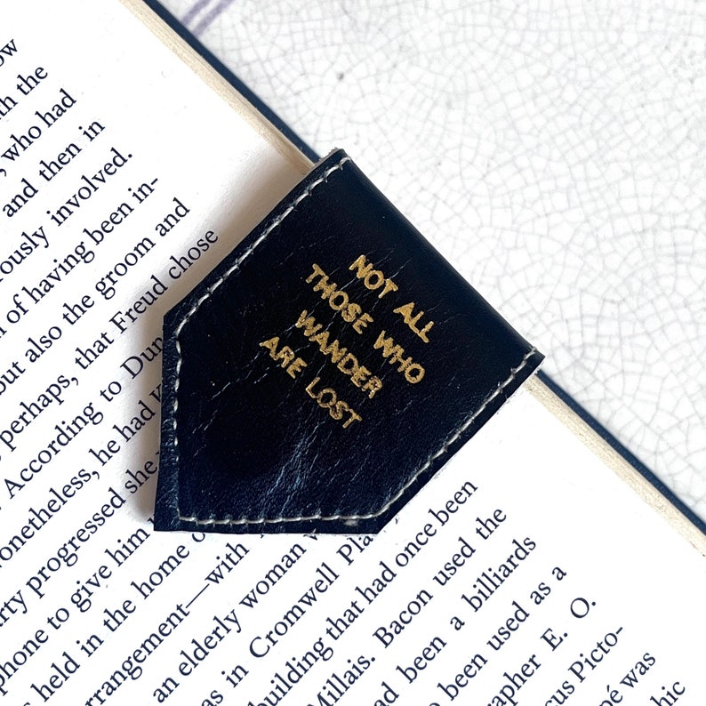 May include: A black leather bookmark with white stitching and gold lettering. The bookmark is in a corner shape and reads "NOT ALL THOSE WHO WANDER ARE LOST". The bookmark is placed on the corner of a book.