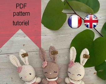 May include: Three crocheted bunny plushies in pink, brown, and white, wearing pink dresses with ruffled edges. The plushies are sitting on a light wood surface.