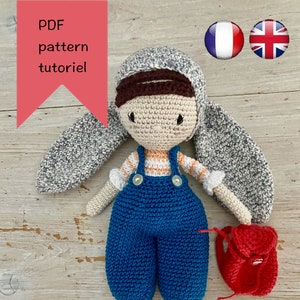 May include: A handmade crochet doll with a gray bunny ear hat, blue overalls, and a small red backpack. The doll has a cream-colored face and limbs, with a striped shirt. The text "PDF pattern tutoriel" is in a red banner.
