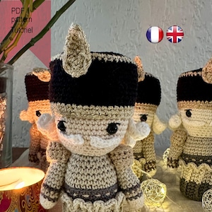 Może przedstawiać: Szydełkowana lalka-żołnierz z czarnym kapeluszem, złotymi ozdobami i białą brodą. Lalka stoi na stole z białym obrusem i światełkami.