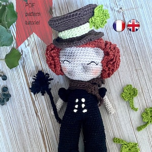 Peut inclure: Une poupée en crochet habillée en ramoneur. La poupée a les cheveux roux, un chapeau brun avec un trèfle vert et une tenue noire et blanche. La poupée tient un balai de ramoneur noir et blanc. Il y a trois décorations en trèfle vert sur la surface en bois.