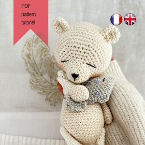 May include: A crocheted white teddy bear holding a smaller crocheted gray and purple fish. The bear has a black nose and closed eyes.