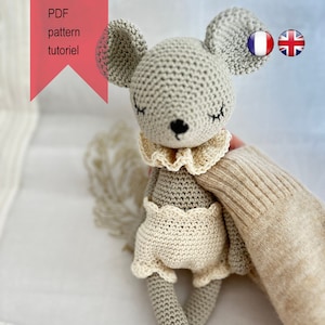 以下が含まれることがあります： 目を閉じたグレーの毛糸で編まれたネズミのぬいぐるみ。白い毛糸で編まれた襟とスカートを着用しています。ぬいぐるみは白と茶色のストライプの表面に座っています。