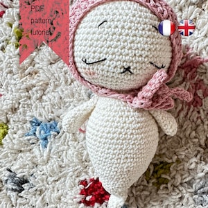 Könnte beinhalten: Ein weißes gehäkeltes Amigurumi-Spielzeug mit einer rosa gehäkelten Mütze und einem Schal. Das Spielzeug hat ein lächelndes Gesicht mit schwarzen Augen und einem gestickten Mund. Das Spielzeug liegt auf einem weißen, flauschigen Teppich mit bunten Akzenten.