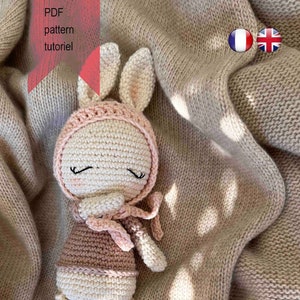 Peut inclure: Un jouet en crochet amigurumi en forme de lapin, réalisé avec du fil rose et blanc. Le lapin porte un chapeau rose et une écharpe rose. Le lapin est couché sur une couverture en tricot beige.