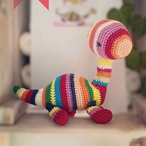May include: A handmade, colorful crochet dinosaur toy. The dinosaur features a long neck and tail, with a striped pattern in various colors including red, yellow, green, and blue. The text "PDF pattern tutoriel" is visible in the image.