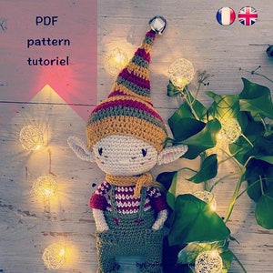 Op de afbeelding: Gehaakte elfpop met een rood, geel en groen gestreepte muts, die een rood-wit gestreept shirt en een groene overall draagt. De pop zit op een houten oppervlak met lichtslingers.