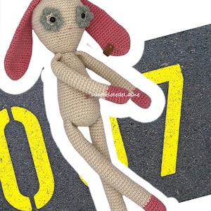 Può includere: Un coniglio di peluche lavorato a uncinetto con orecchie rosa e occhi a forma di fiore verde. Il coniglio è in piedi su una superficie grigia con segni gialli.