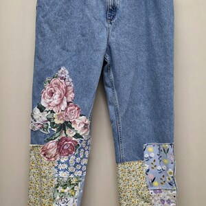 May include: Light blue denim jeans with floral patchwork details. The jeans feature a large floral appliqué on the left leg and patchwork on the cuffs. The patchwork includes various floral patterns in pink, yellow, and blue.