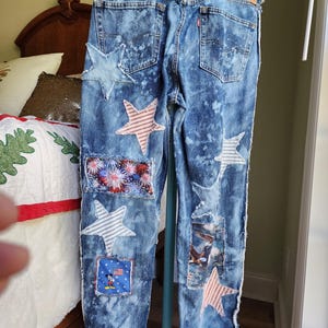 May include: Blue denim jeans with star and patriotic-themed patches. The jeans have a light blue wash with white accents. Patches include stars, fireworks, an eagle, and a Mickey Mouse graphic. The bottom hem features a blue band with white stars.