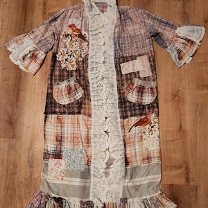 May include: A patchwork duster coat with a mix of plaid and floral patterns in shades of brown, red, and cream. The coat features lace trim, ruffled sleeves, and decorative bird and floral appliqués. The coat has two pockets and a ruffled hem.