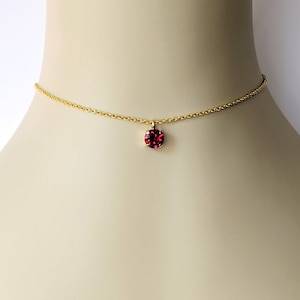 May include: A delicate gold chain necklace with a small, round, dark pink gemstone pendant.