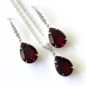 May include: A set of jewellery featuring a teardrop-shaped pendant and earrings with a deep red gemstone and a silver chain with small clear stones.
