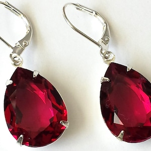Könnte beinhalten: Ein Paar silberne Ohrhänger mit tropfenförmigen roten Kristallsteinen.