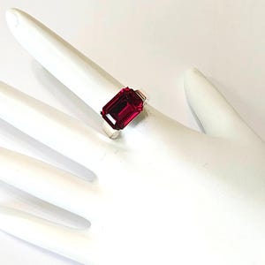 May include: A silver ring featuring a large, rectangular, deep red gemstone. The ring is set on a white hand, showcasing the vibrant color and design of the jewelry. The gemstone's cut and clarity are prominent.