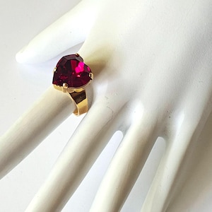 May include: A gold-toned ring featuring a large, heart-shaped, ruby-red gemstone. The ring is set on a white surface, showcasing the vibrant color and design. The ring's band is a simple, classic style.