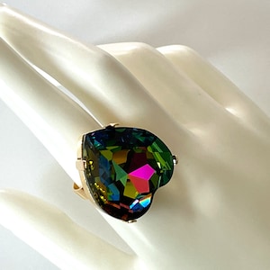 May include: A gold-toned ring featuring a large, heart-shaped gemstone. The gemstone displays a vibrant array of colors, including green, blue, and pink, creating a striking iridescent effect. The ring's setting is simple, allowing the gemstone to be the focal point.