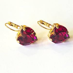May include: A pair of heart-shaped earrings with a deep red gemstone set in gold-colored metal. The earrings have a lever-back closure and are displayed on a white surface. The gemstones are faceted, catching the light.