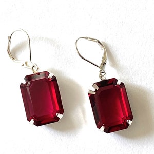 May include: A pair of rectangular, ruby-red earrings with silver-colored lever-back closures. The earrings have a faceted appearance and are set in a silver-toned metal. The earrings are displayed against a white background.