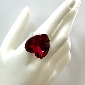 May include: A silver-toned ring featuring a large, heart-shaped, faceted, deep red gemstone. The gemstone is the focal point, catching the light and reflecting a vibrant color. The ring's band is simple, allowing the heart-shaped stone to stand out.