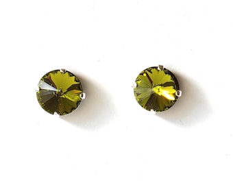 May include: Two olive-green crystal stud earrings with silver-colored prongs. The round crystals have multiple facets that reflect light. The earrings are set against a white background, highlighting their color and design.