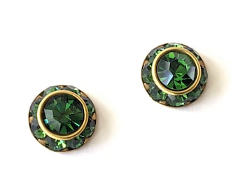 May include: A pair of gold-tone earrings with green crystal stones. The earrings are round and have a bezel setting.