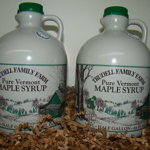 May include: Two white ceramic jugs with green lids, filled with maple syrup. The jugs are labeled "Trudell Family Farm Pure Vermont Maple Syrup" and have a picture of a farmhouse and trees. Each jug holds a half gallon (64 fl oz).