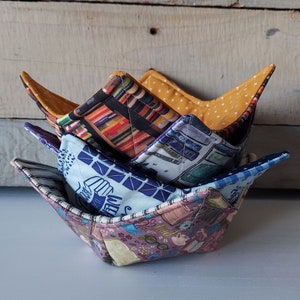 May include: Three fabric bowls stacked on top of each other. The top bowl is yellow with white polka dots, the middle bowl is blue with a book pattern, and the bottom bowl is purple with a book and glasses pattern. The bowls are all quilted and have a scalloped edge.