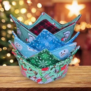 May include: A stack of four fabric bowl cozies in a variety of holiday patterns, including a snowman, a penguin, and a snowflake design. The cozies are folded into a crown shape and are made of quilted fabric.