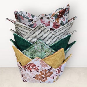 May include: A stack of handmade bowl cozies in various floral and patterned fabrics. The cozies feature a variety of colors, including white, green, yellow, and brown. Each cozy is designed to hold a bowl and protect hands from heat.