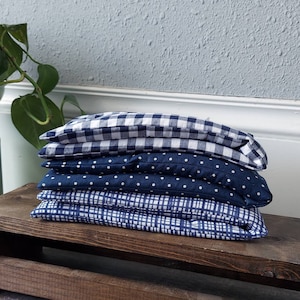 May include: Four blue and white patterned eye pillows stacked on a wooden shelf. The pillows are made of fabric with different patterns, including gingham, polka dots, and stripes.