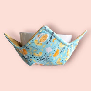 May include: A blue and yellow bowl cozy with a pattern of macaroni and cheese. The text on the cozy reads "You're so cheesy! I love you dearly! Macaroni!"