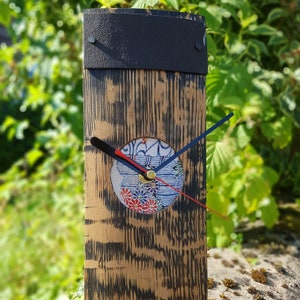 May include: A wooden clock with a black metal top and a decorative clock face. The clock face features a floral design with red, blue, and white accents. The clock hands are black, blue, and red.