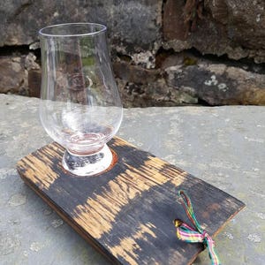 May include: A small, clear glass sits on a wooden coaster with a dark brown finish. The coaster has a hole in the center and is tied with a colorful ribbon.