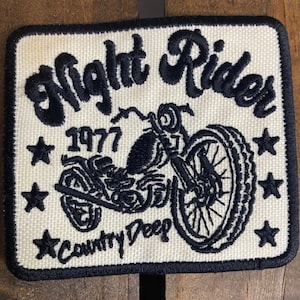 May include: A white embroidered patch with black stitching featuring a motorcycle, the text "Night Rider", "1977", and "Country Deep".