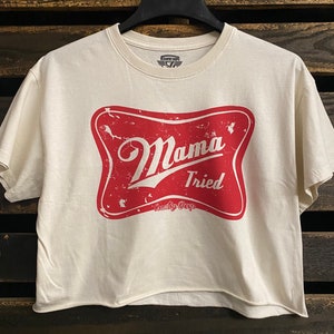 Op de afbeelding: Een wit gecropped t-shirt met een rode, verweerde grafische afbeelding van een ster met de tekst "Mama Tried" en "Country Deep" erin.