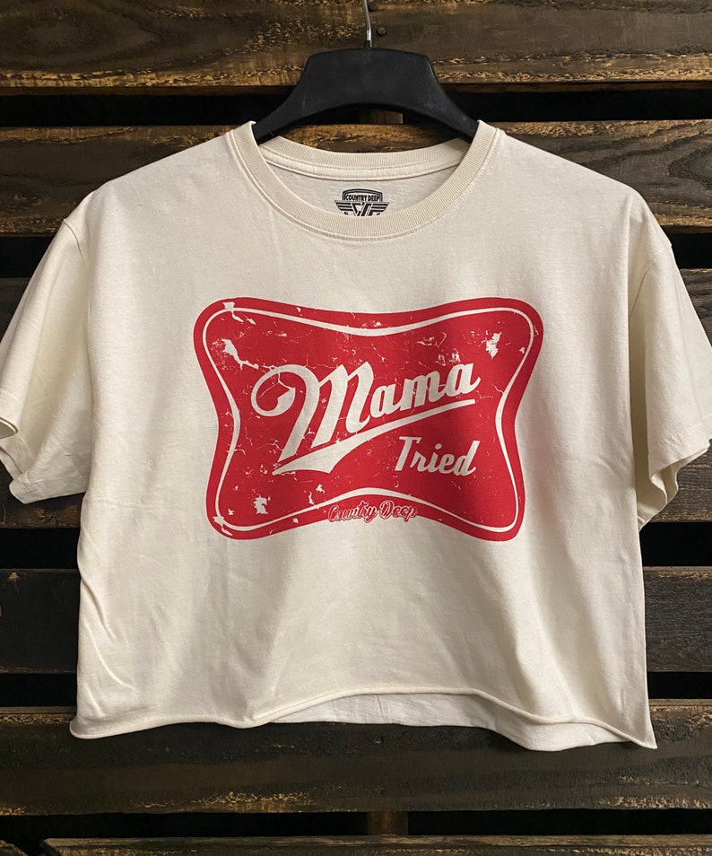 Op de afbeelding: Een wit gecropped t-shirt met een rode, verweerde grafische afbeelding van een ster met de tekst "Mama Tried" en "Country Deep" erin.