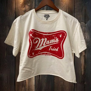 Op de afbeelding: Een wit gecropped t-shirt met een rood, verweerd logo met de tekst "Mama Tried" en daaronder de woorden "Country Deep".