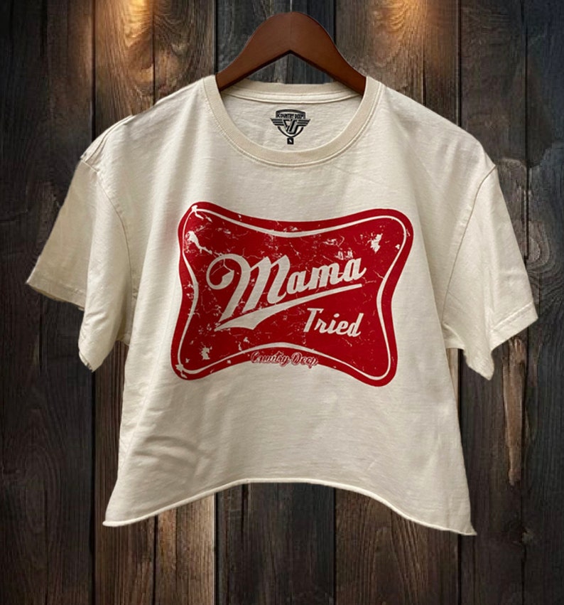 Op de afbeelding: Een wit gecropped t-shirt met een rood, verweerd logo met de tekst "Mama Tried" en daaronder de woorden "Country Deep".